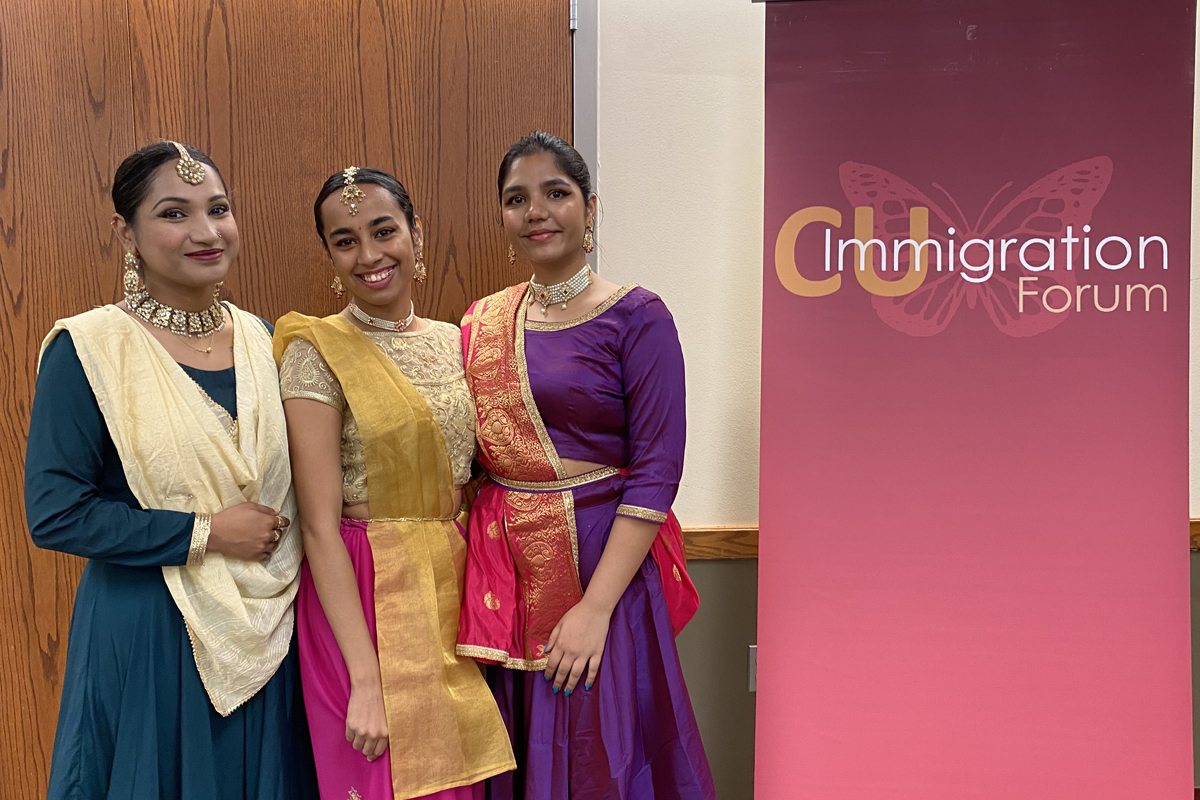 Three smiling dancers in colorful dresses standing next to a banner that says "CU Immigration Forum"