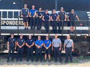 Group photo of the Champaign firefighters who participated in the training exercise. They stand in two rows. One row of 7 stands on the ground. A second row of 7 stands directly above them on the side of the train.