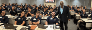 Image of Tracy Parsons (center-right, standing) with a class full of cadets at the Police Training Institute. He's standing next to a student facing the camera for a group/classroom photo.